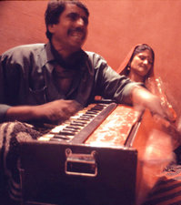 Man playing a harmonium.  He is pumping the bellows of the harmonium with one hand and playing it with the other.